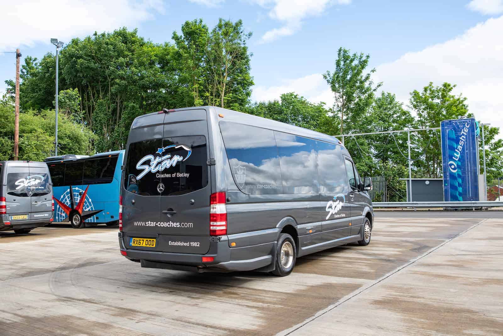 A grey "Star Coaches of Batley" 16 seater minibus hire with driver parked on a paved area with trees in the background.