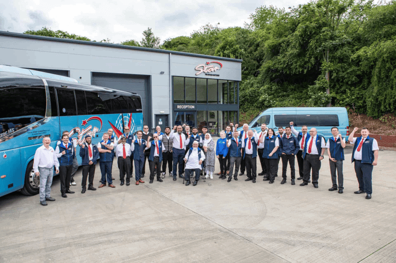 Group of employees in smart uniforms gather outside a building marked 'Star' next to a blue bus and van. The atmosphere is cheerful and team-oriented — perfect time to Book a Coach in Advance.
