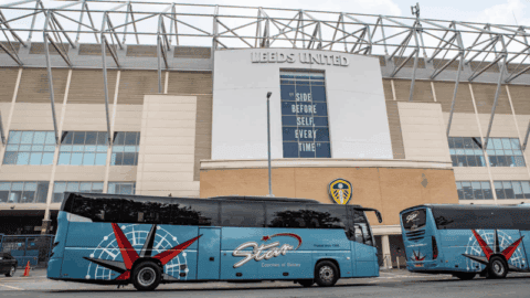 Two blue coaches parked outside a stadium with 'Leeds United' signage. The wall displays the motto 'Side Before Self Every Time' — a great reminder to Book a Coach in Advance for matchday travel.