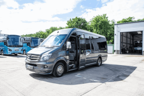 A sleek gray minibus with open doors is parked in a sunny lot beside several blue coaches and a garage. Lush green trees in the background — a smart moment to Book a Coach in Advance.