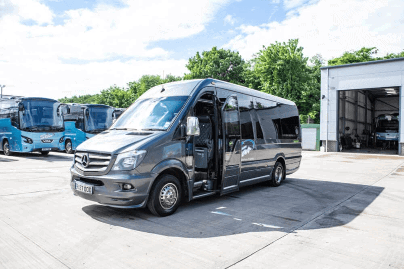 A sleek gray minibus with open doors is parked in a sunny lot beside several blue coaches and a garage. Lush green trees in the background — a smart moment to Book a Coach in Advance.