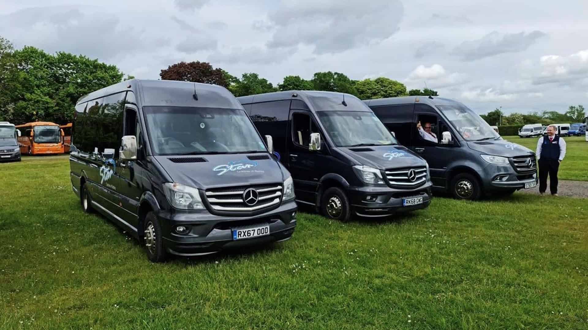 Three dark gray Mercedes-Benz minibuses parked on grass with trees and a cloudy sky in the background.