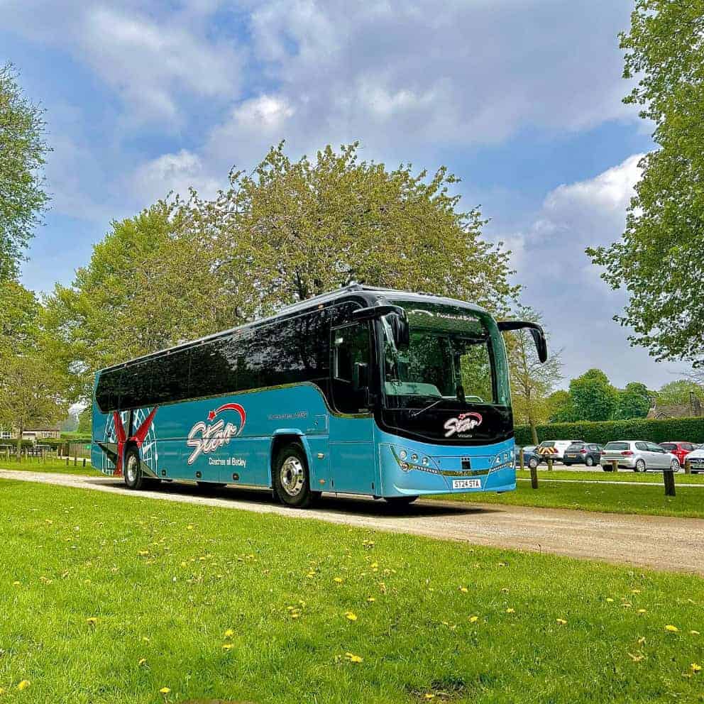 A blue tour bus on a park road during a UK road trip, surrounded by greenery and trees under a cloudy sky.