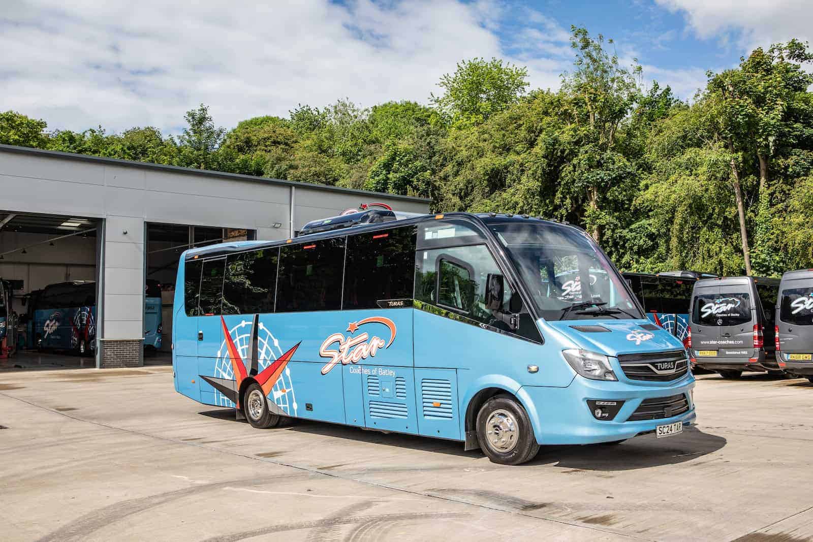 Light blue bus parked near a garage ready for a UK road trip on a sunny day, surrounded by lush greenery and other buses.