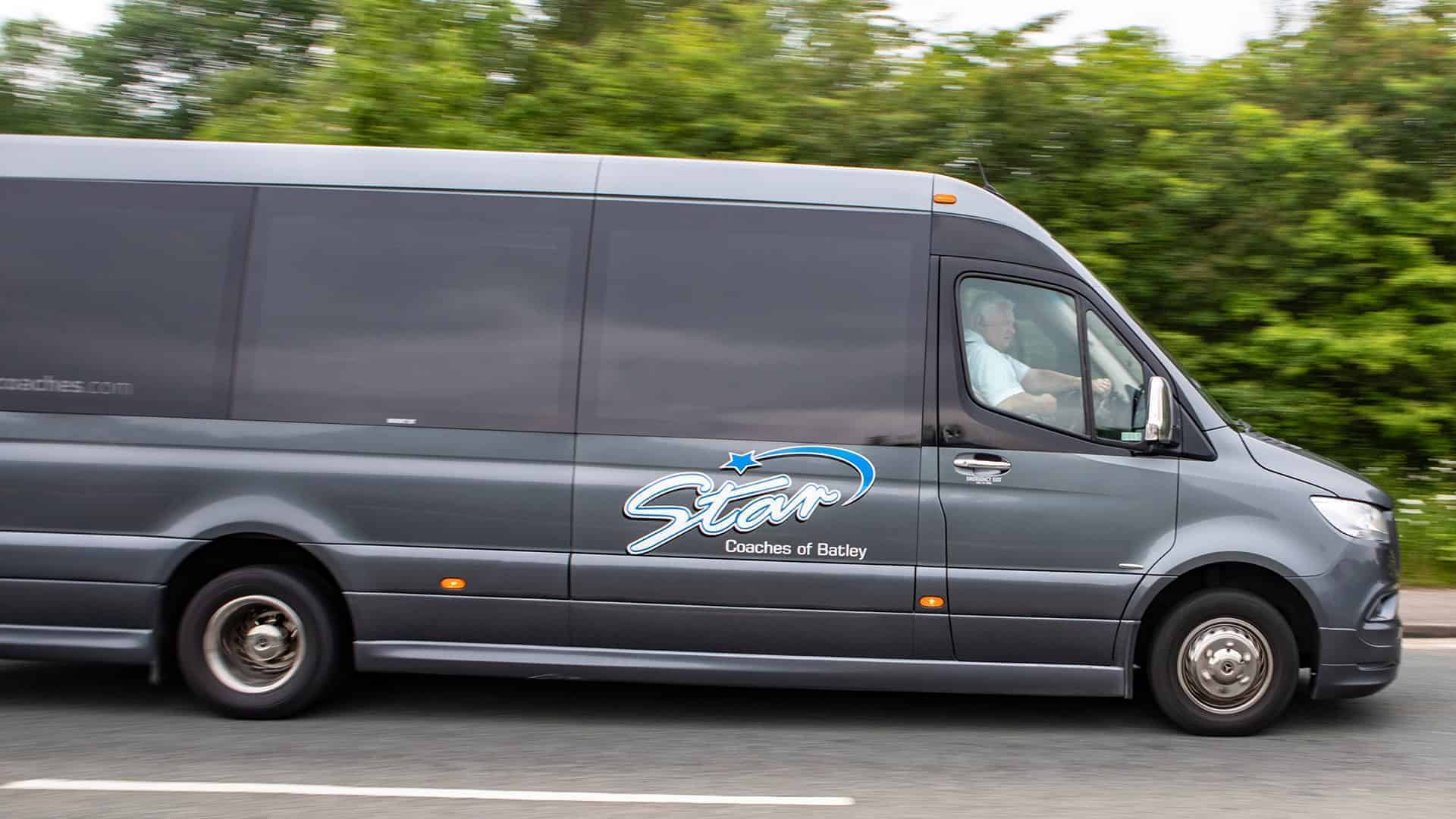 A dark gray 16 Seater Minibus Price, labeled "Star Coaches of Batley" on a road with a blurred green tree background.