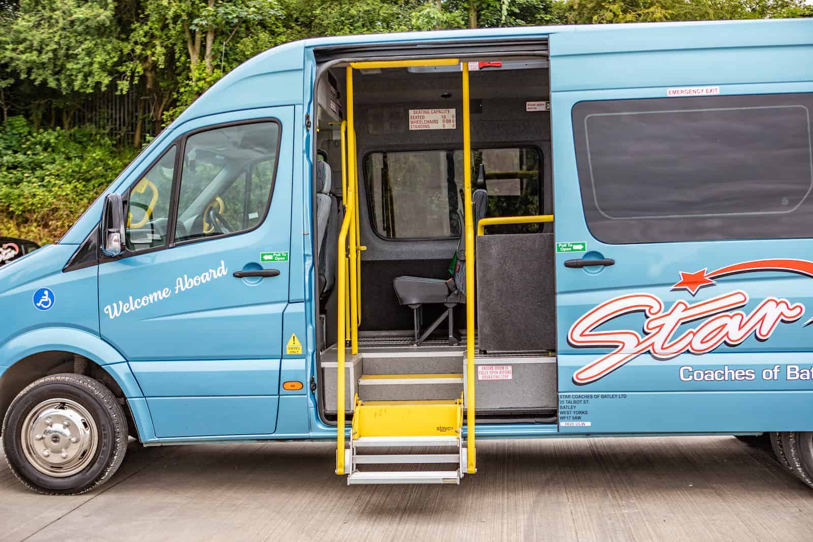Accessible minibus 2 A blue accessible minibus with an open side door, featuring a yellow handrail and steps, parked on a concrete surface with greenery in the background.