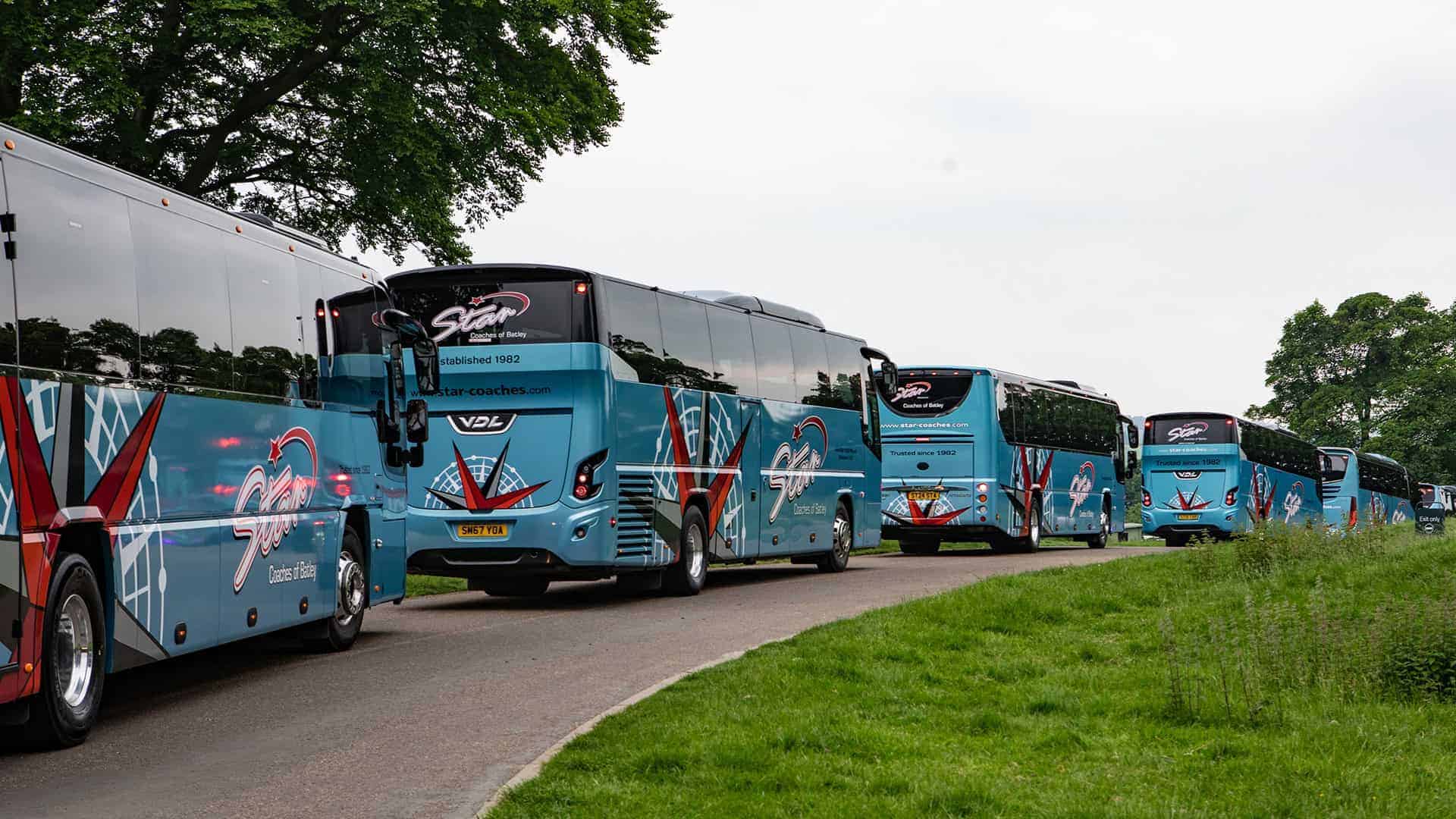 Coach Hire fare 2 Blue tour buses with red and white accents drive on a winding road surrounded by lush green trees and grass.