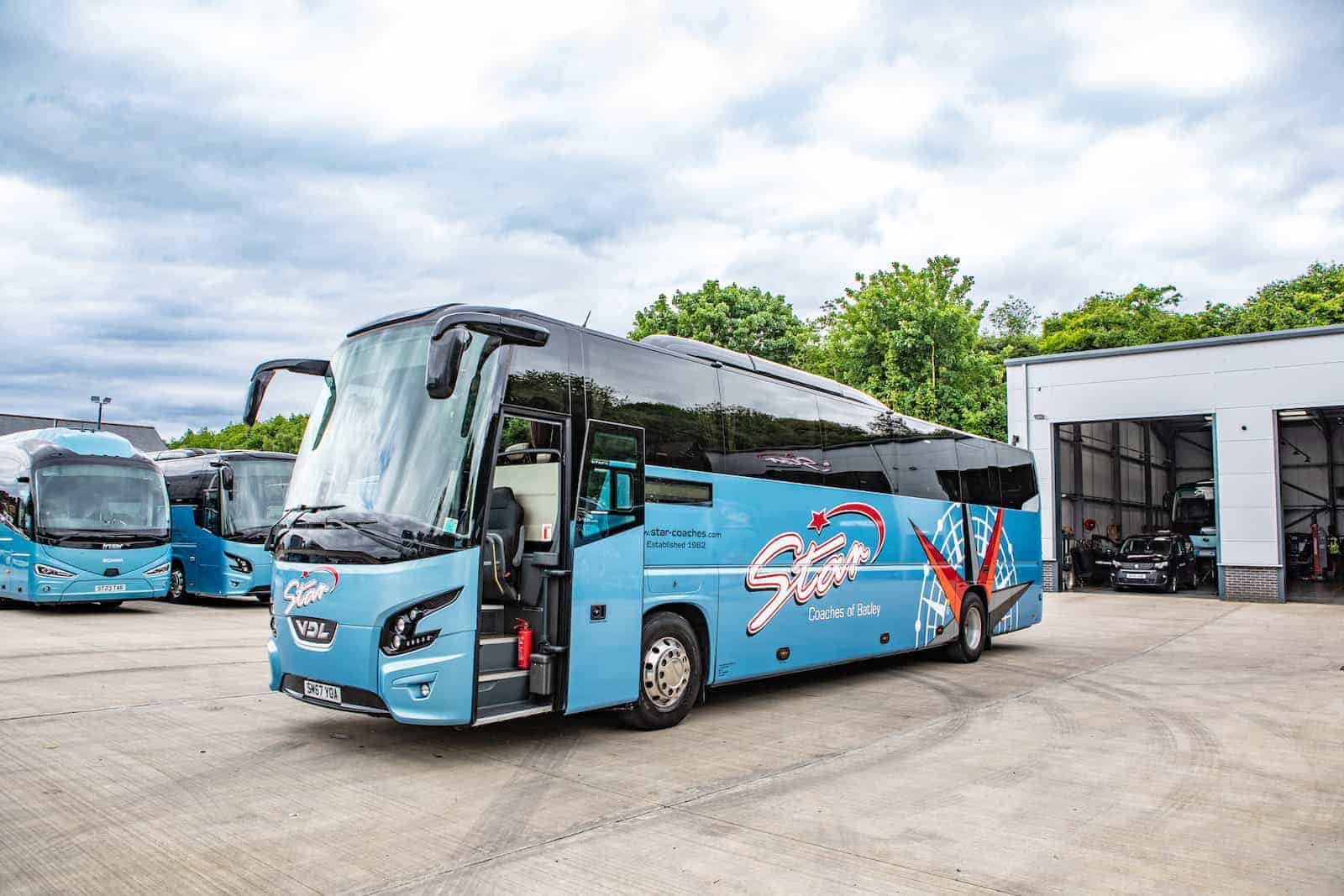 A modern blue bus with a “Star” logo parked outside a garage on a cloudy day, surrounded by trees and other buses.