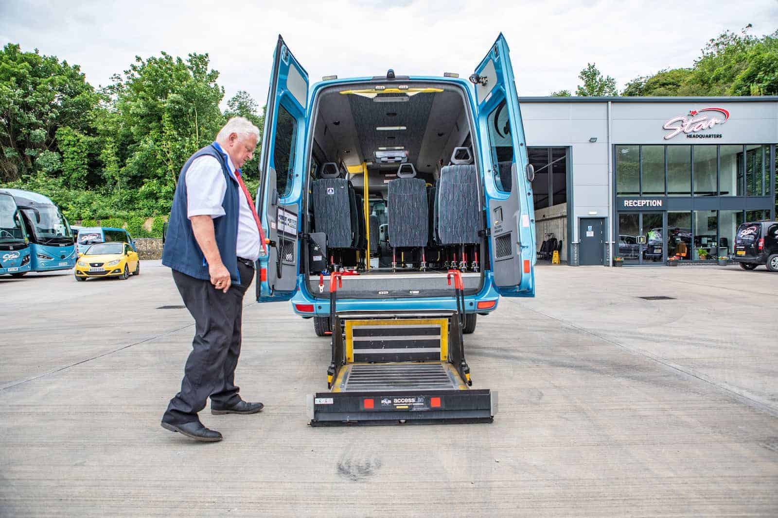 Man standing beside an open blue van with a wheelchair lift extended, parked near a building marked “reception.”