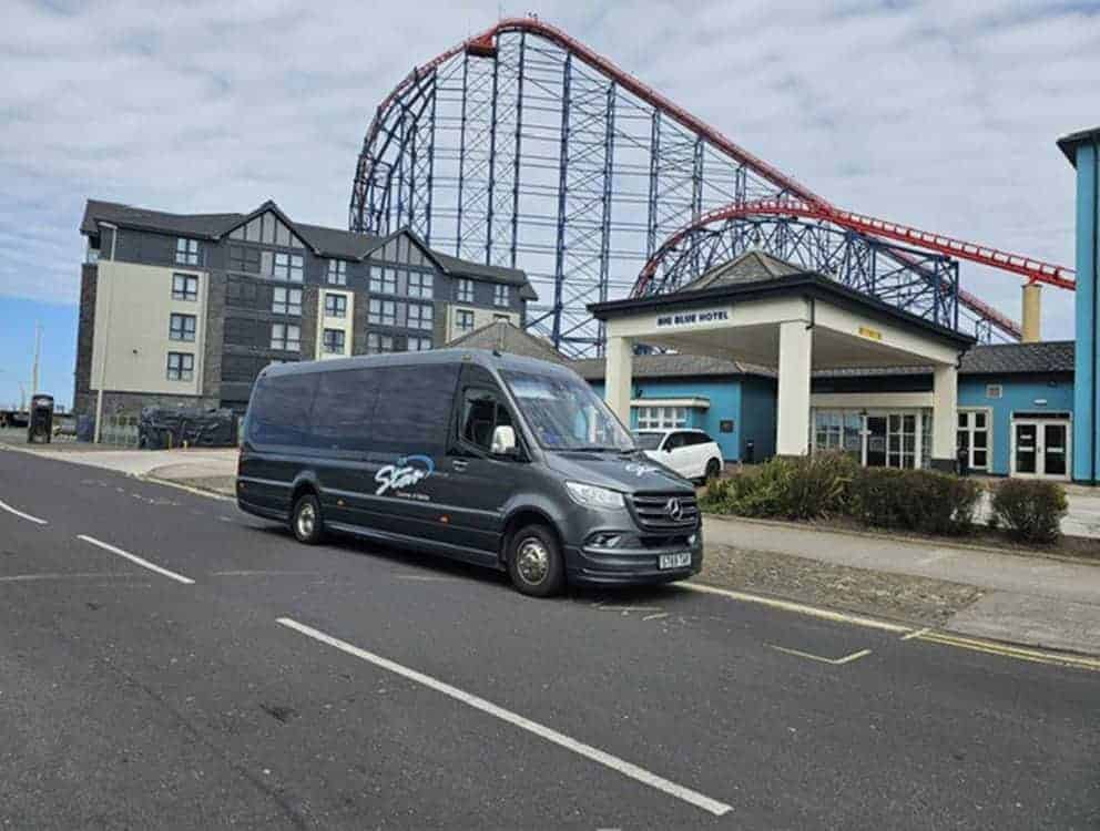 A grey van parked beside a modern hotel with a red and blue roller coaster rising behind it under a bright, clear sky.