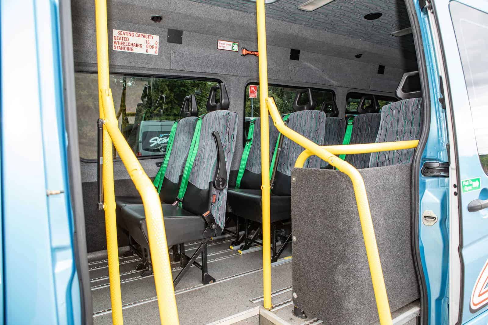 Interior of blue minibus with gray seats, green seat belts, yellow rails and capacity sign, showing trust Coach Hire PSVAR UK.

