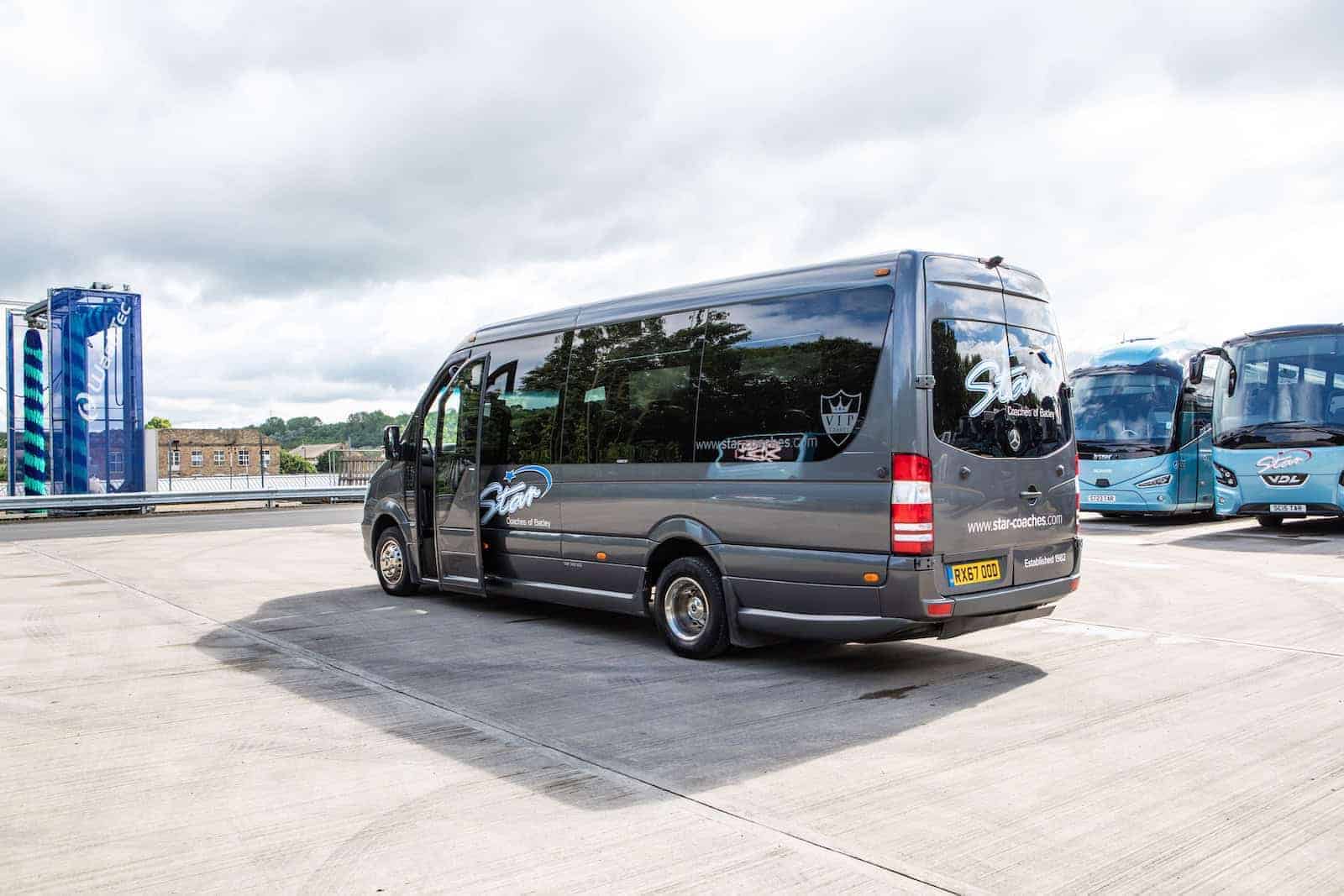Gray minibus parked on concrete lot with blue coaches behind under cloudy sky, showing Coach hire pre-trip checklist UK.
