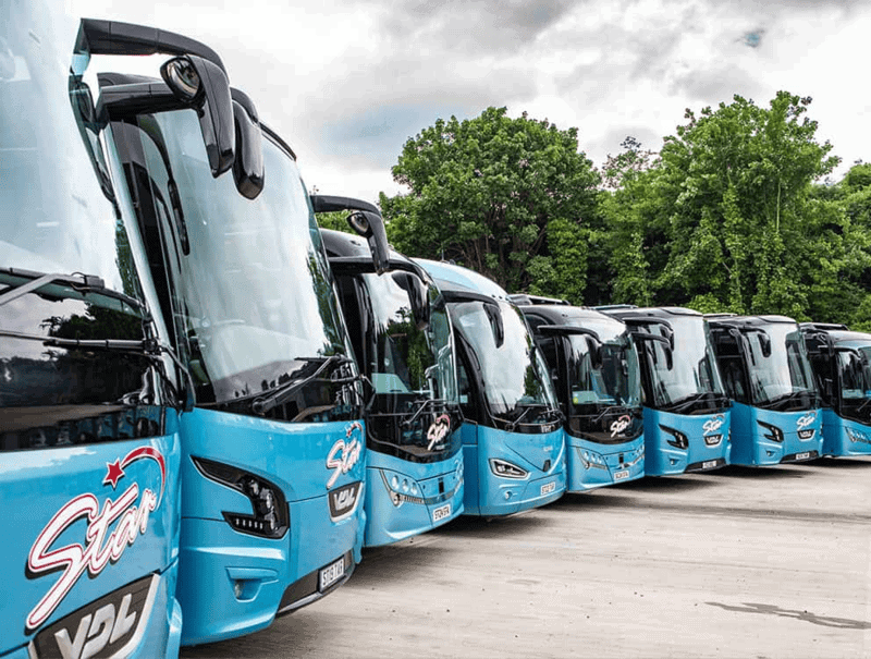 Blue tour buses parked in a row on a paved lot with green trees and cloudy sky, showing wedding coach hire multiple pick-ups UK.