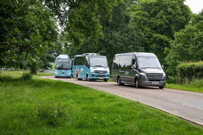 Three minibuses driving in a line on a tree-lined park road with bright greenery and overcast sky, Winter Coach Travel UK.
