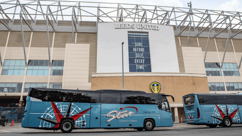 Two blue coaches with red accents parked outside Leeds United stadium, showing a professional athletic vibe, highlighting how coach operators ensure safety vs aggregators through well-maintained, branded vehicles.