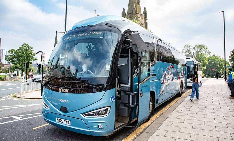 A sleek blue Scania tour bus parked on a city street with its door open as people board, and a historic church visible under a partly cloudy sky.